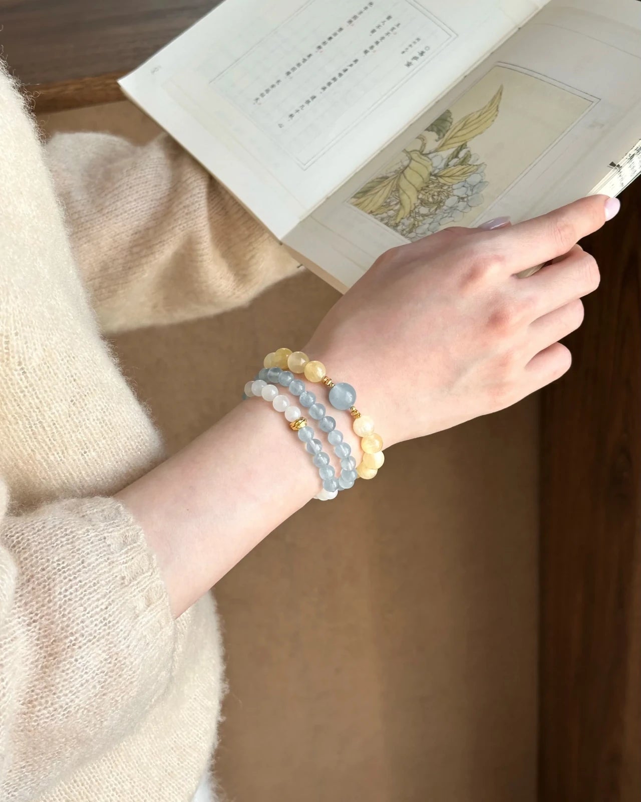 Lifestyle wrist shot—aquamarine and white moonstone (moon stone) 6mm bracelet with gold-plated accents, stacked while reading.