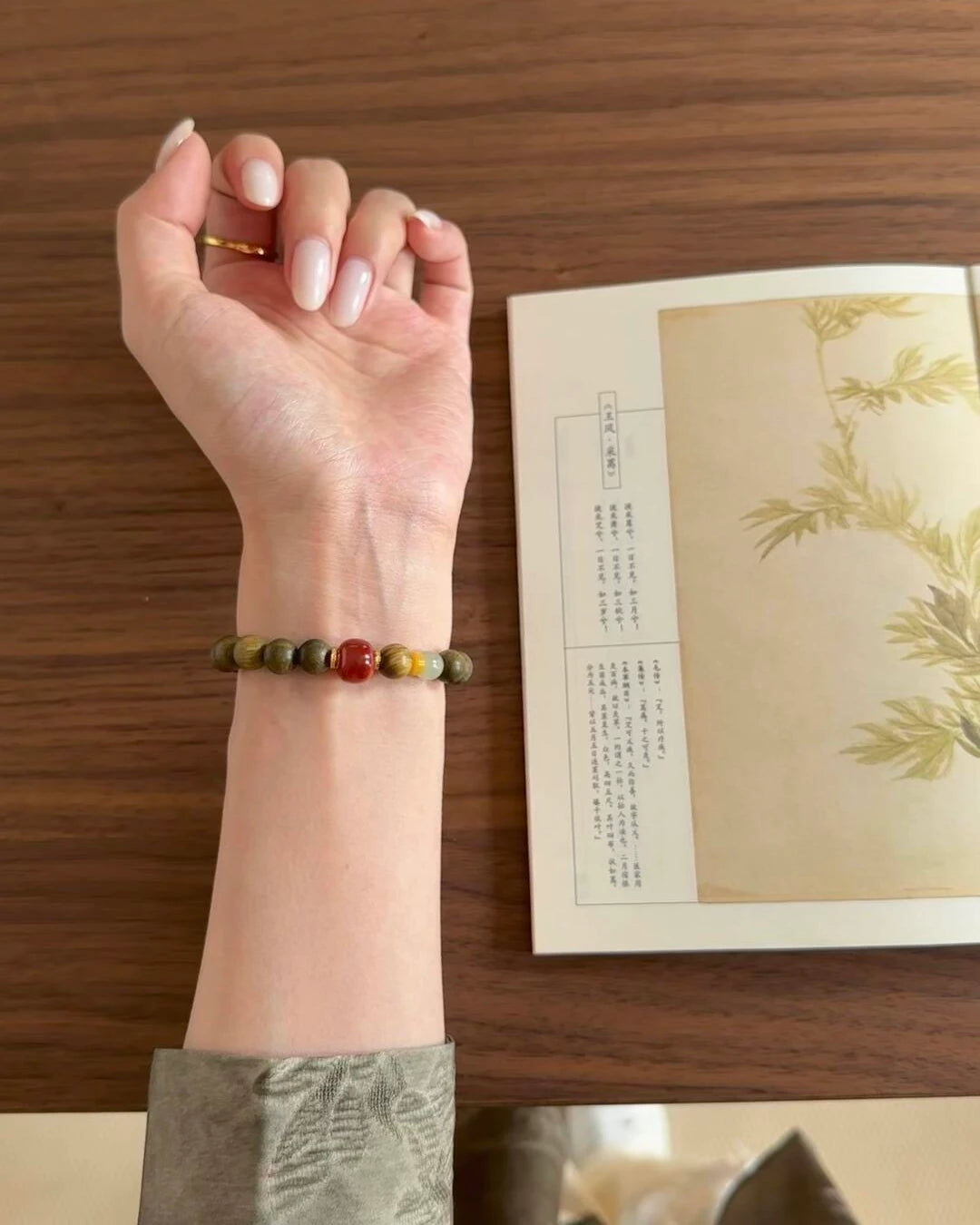 Vital Harmony Bracelet on wrist beside an open book, showing sandalwood beads with red agate, amber, and jade.