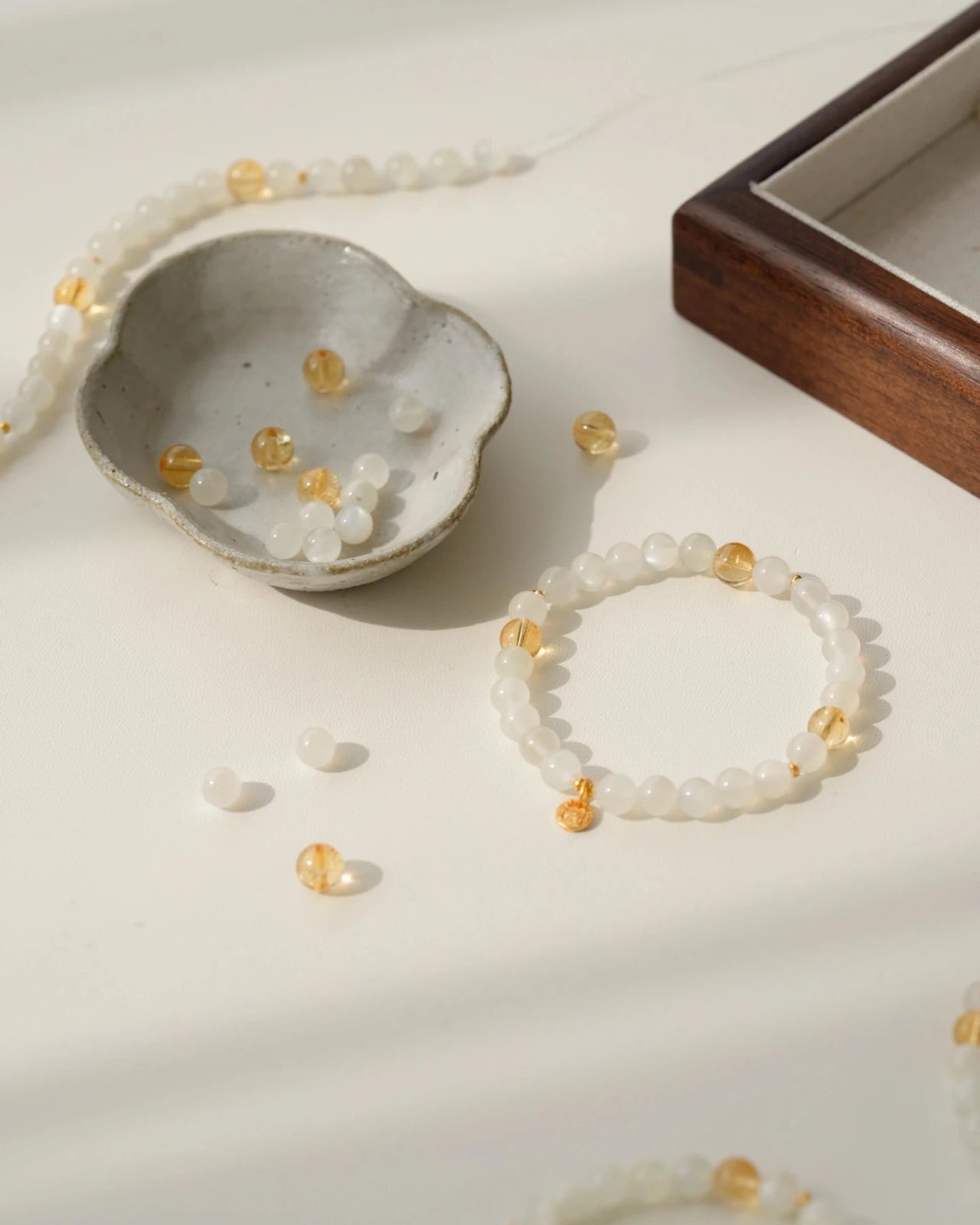Flat lay in natural light—white moonstone bracelet with citrine accents and a small gold-plated S925 charm, styled with loose beads and a ceramic dish.