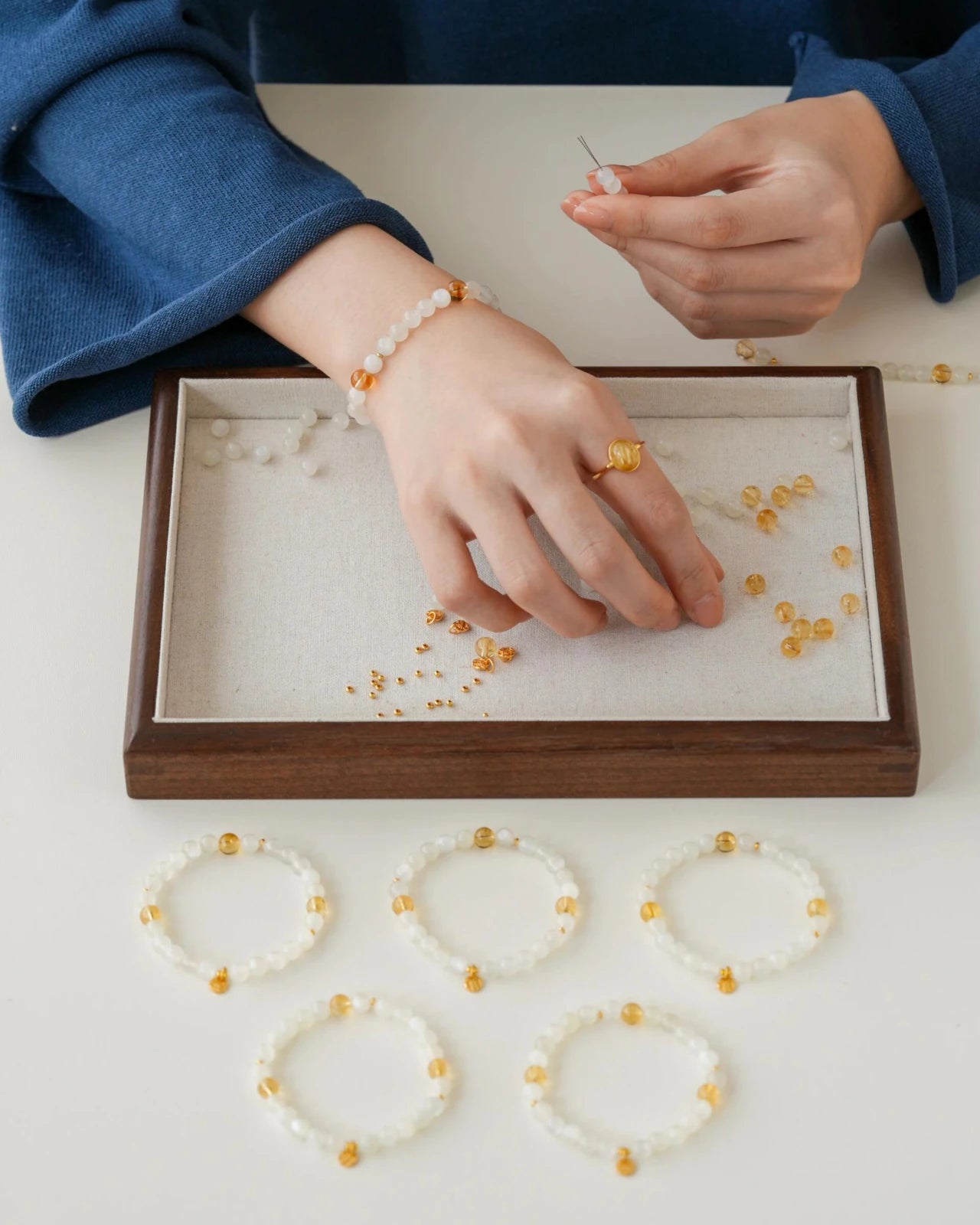 Top-down view—white moonstone bracelet with citrine accents and a small gold-plated S925 charm; wrist shown beside multiple finished pieces arranged on a bead tray.