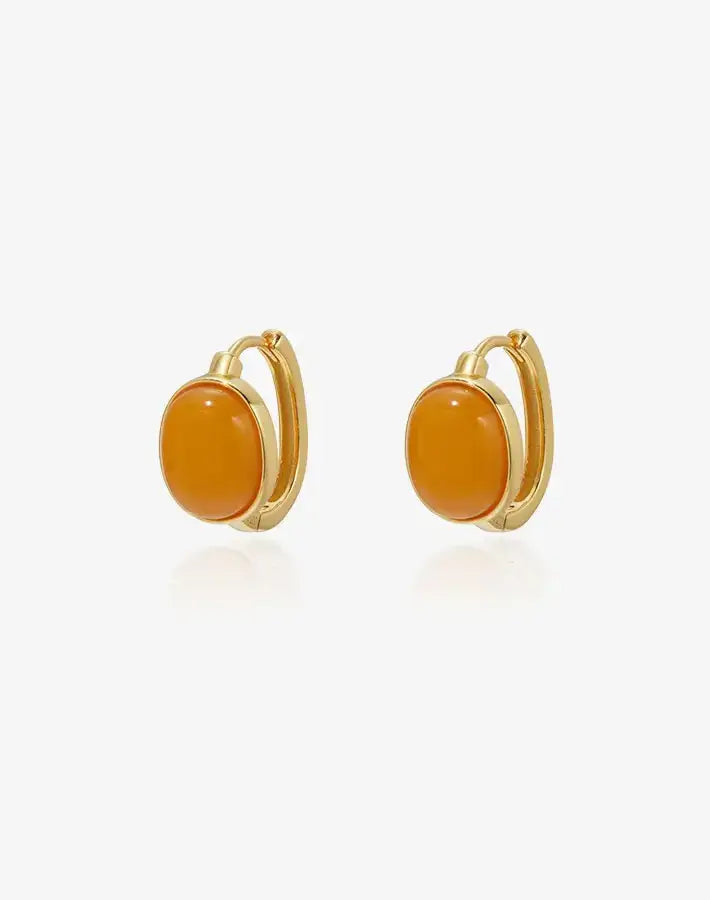 Flat lay: Golden Ember earrings with oval amber cabochons in gold-plated S925 silver huggie hoops on a gray background.