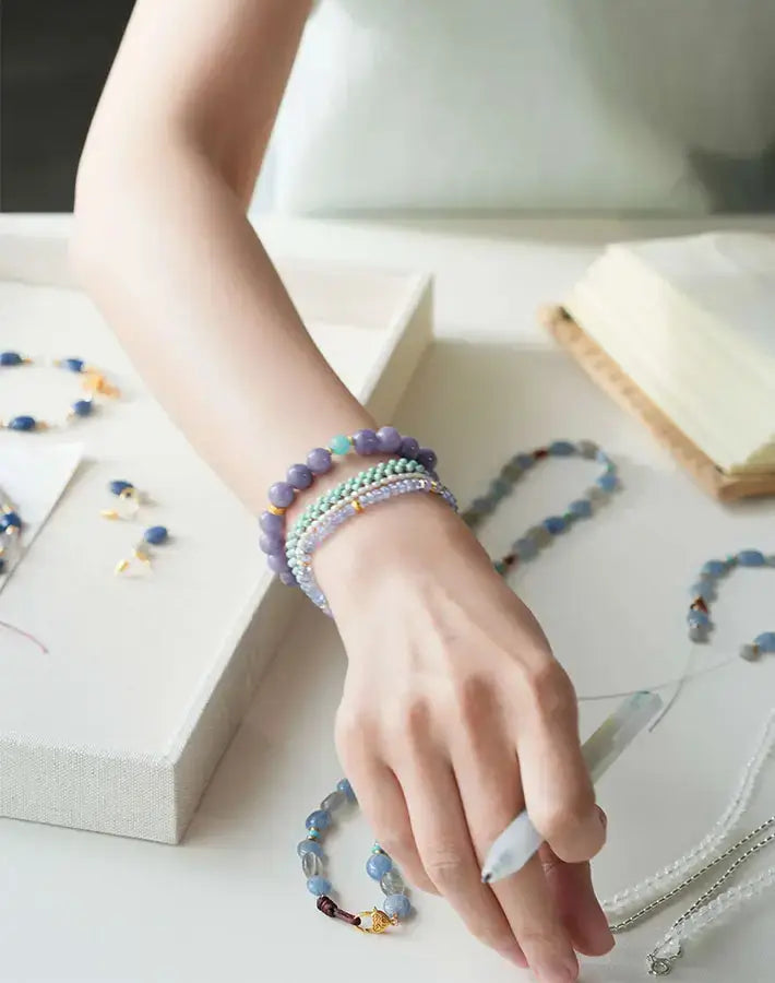 Arm reaching across a desk showcasing the Lavender Tide Serenity bracelet—purple chalcedony and amazonite beads with gold-plated S925 silver spacers—alongside other gemstone accessories.