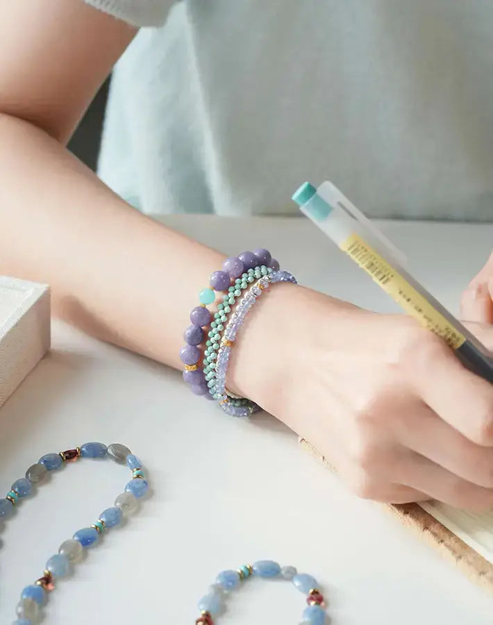 Wrist adorned with the Lavender Tide Serenity bracelet of purple chalcedony and amazonite beads with gold-plated S925 silver spacers, hand writing in a notebook.