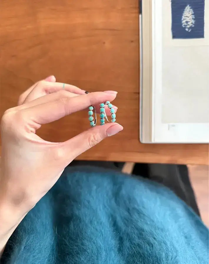 Hand holding three Skyward Rings upright, showing rows of cushion turquoise cabochons on sterling-silver bands beside an open book.
