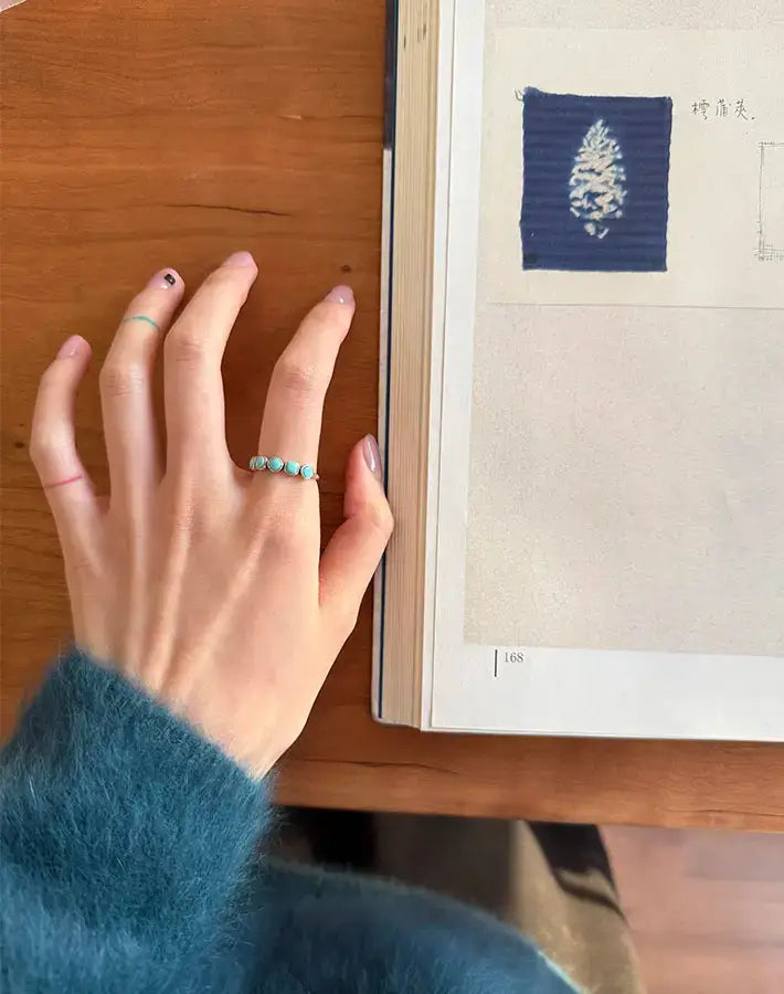Top-down desk scene: hand resting near open book wearing Skyward Ring with five square-cut turquoise stones on sterling-silver band.
