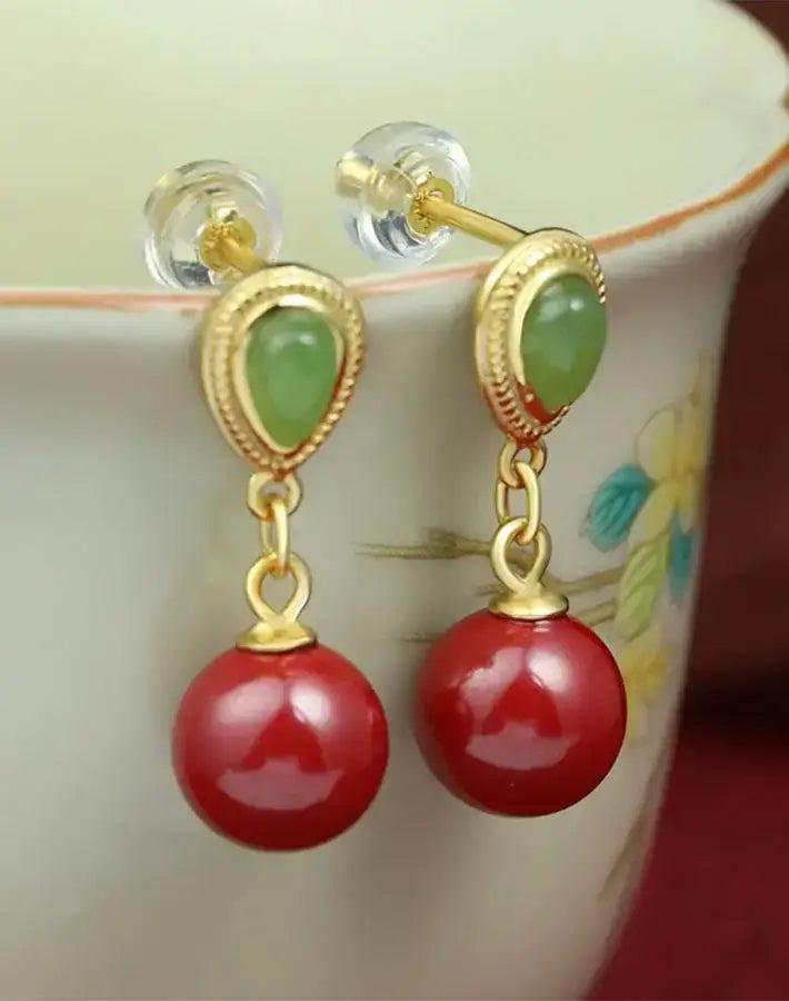 Vital Grace earrings—nephrite studs and cinnabar drops in gold-plated S925 silver—hanging from a floral porcelain cup rim.