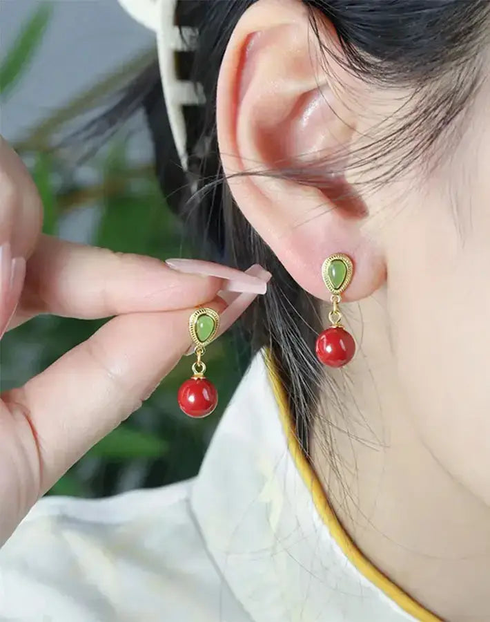Close-up of ear wearing Vital Grace earring—nephrite stud, cinnabar drop and gold-plated S925 silver—while hand shows the second piece.